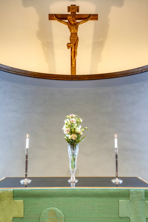 Kuvassa on Töölön kirkon alttari, joka on puettu vihreään alttarivaatteeseen. Alttarilla on kaksi kynttilää ja kukkia maljakossa. Alttarin yläpuolella puusta veistetty kuva ristiinnaulitusta Jeesuksesta.