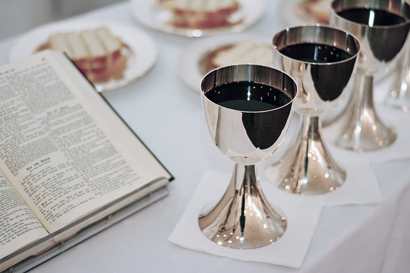 Kuvassa avonainen Raamattu, hopeisia pikareita, joissa on viiniä ja ehtoollisleipä.