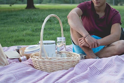 Tunnelmakuva. Kuvassa piknik-peitto, eväitä ja kesäinen nurmikko. Mies istuu piknikillä shortseissa ja t-paidassa.