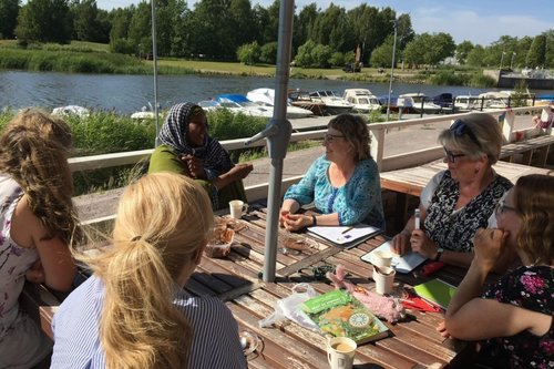 Suomalaisia ja maahanmuuttajia kahvittelemassa kesällä terassilla veden äärellä kielikahvilassa.