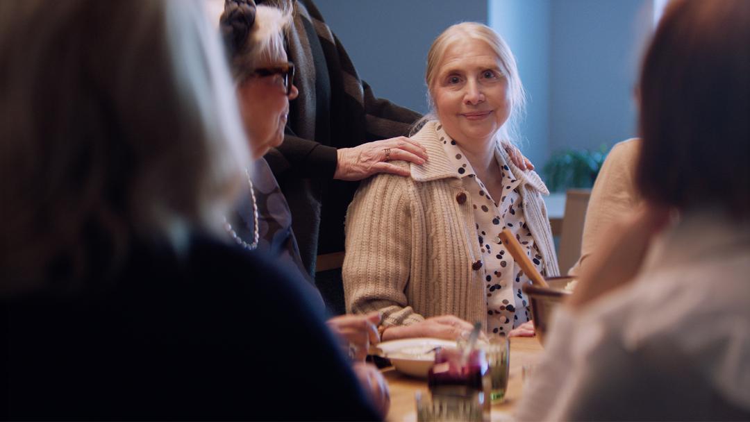 Nainen istuu ruokapöydässä toisten seurassa. Hän hymyilee lempeästi kameraan.
